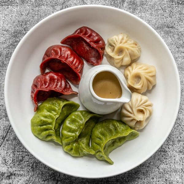 A white plate holds red, green, and white Nepali momo dumplings with a small pitcher of sauce.