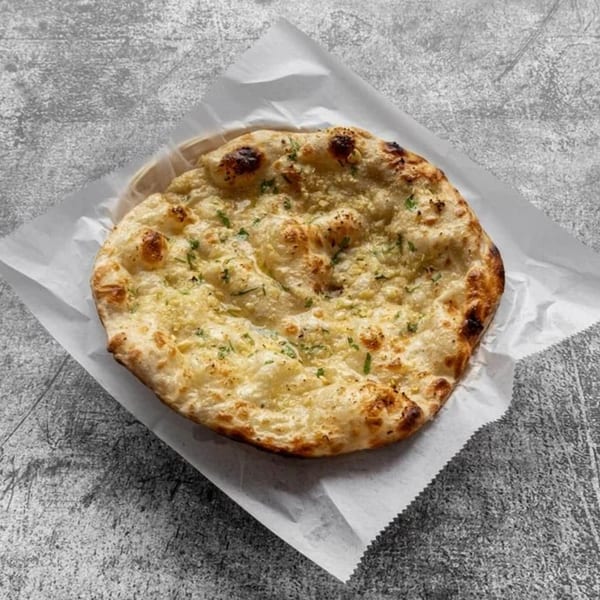 A round, golden-brown garlic naan bread, sprinkled with herbs, rests on white parchment paper.
