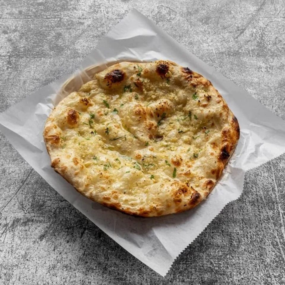 A round, golden-brown garlic naan bread, sprinkled with herbs, rests on white parchment paper.