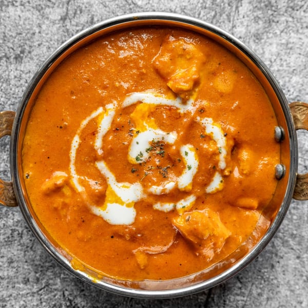 A close-up of Nepali butter chicken in a metal bowl, garnished with cream swirls and herbs.